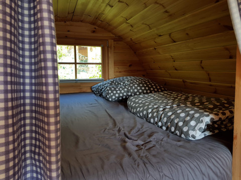 Campingpark im Bergischen Land Kleines Holzpod mit gemütlicher Schlafnische, Fensterblick und kariertem Vorhang für naturnahes Camping.