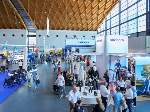 Ludzie w hali wystawowej z wózkami inwalidzkimi, produkty Meyra i Ottobock, jasne otoczenie.