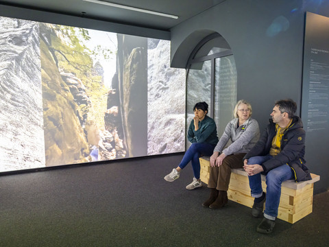 Drei Personen sitzen auf einer Holzbank in einem Museum, betrachten eine große Projektion von Felsen und Bäumen. Die Atmosphäre ist ruhig und nachdenklich.