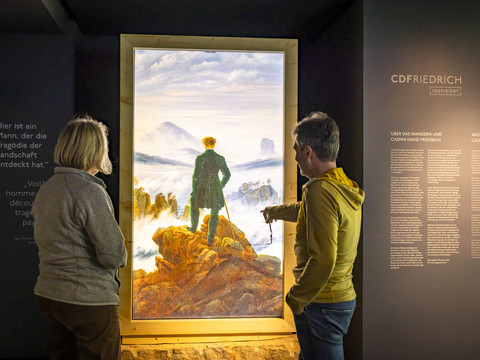 Zwei Personen betrachten ein beleuchtetes Gemälde von Caspar David Friedrich in einer Ausstellung, umgeben von erklärenden Texten an der Wand.