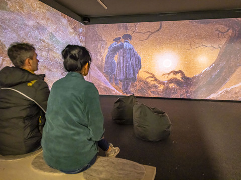 Mediale Ausstellung „CDFriedrich inspiriert“ Zwei Personen sitzen in einem dunklen Raum und betrachten eine große Projektion eines Gemäldes mit zwei Figuren vor einem Sonnenuntergang.