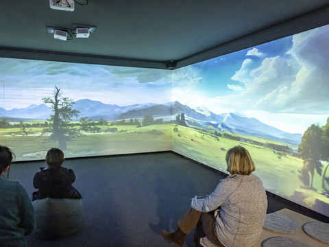 Drei Personen sitzen in einem dunklen Raum und betrachten eine Projektion einer Landschaft mit Bergen, Bäumen und blauem Himmel auf den Wänden.