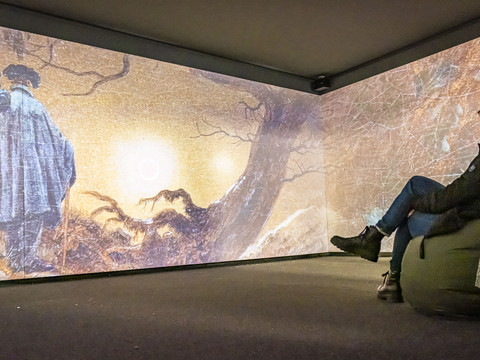 Mediale Ausstellung "CDFriedrich inspiriert“ Eine Person sitzt auf einem Sitzsack in einem Raum mit großflächiger Projektion eines Gemäldes, das zwei Männer in Mänteln zeigt.