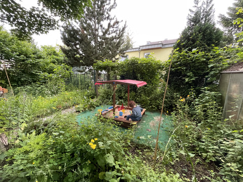 Sandkasten Ein Kind spielt im grünen Garten in einem kleinen Sandkasten mit rotem Dach, umgeben von üppiger Natur.