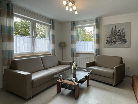 Wohnzimmer Fewo grün Helles Wohnzimmer mit zwei beigen Ledersofas, Glastisch und gemaltem Stadtbild an der Wand.