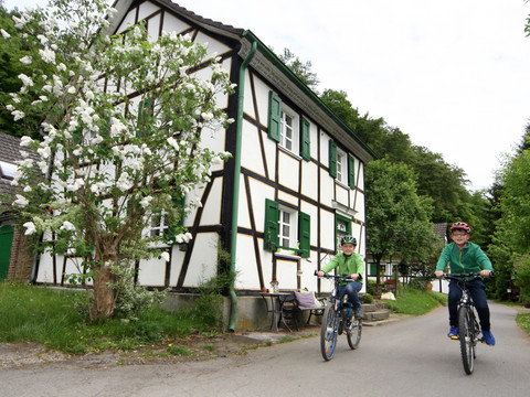 Dhünn-Runde Radfahrer vor einem traditionellen Fachwerkhaus auf einer ländlichen Straße im Grünen.