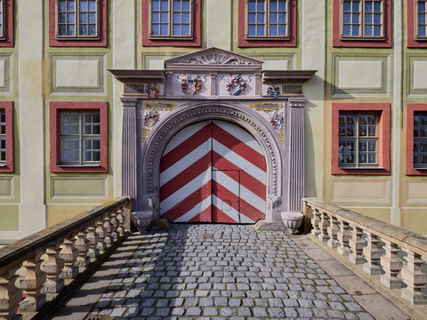 Eingang des Schloss Weesenstein mit pastellgelber Fassade und roten Fensterrahmen, verziertem Torbogen und rot-weiß gestreiftem Holztor, gepflasterter Weg davor.Entrance to Weesenstein Castle with pastel yellow façade and red window frames, decorated archway and red and white striped wooden gate, paved path in front.Vchod do hradu Weesenstein s pastelově žlutou fasádou a červenými okenními rámy, zdobeným obloukem a červenobíle pruhovanou dřevěnou bránou, před ní dlážděná cesta.Wejście do zamku Weesenstein z pastelowo żółtą fasadą i czerwonymi obramowaniami okien, zdobionym łukiem i drewnianą bramą w czerwono-białe pasy, brukowana ścieżka przed wejściem.Ingang van kasteel Weesenstein met pastelgele gevel en rode kozijnen, versierde boog en rood-wit gestreepte houten poort, geplaveid pad ervoor.Ingresso del castello di Weesenstein con facciata giallo pastello e cornici rosse alle finestre, arco decorato e cancello in legno a strisce bianche e rosse, sentiero lastricato sul davanti.