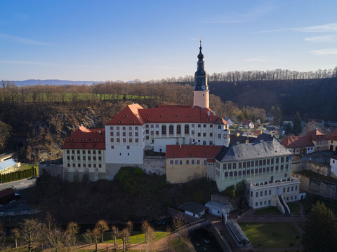 Luftaufnahme von Schloss Weesenstein mit roten Dächern und einem hohen Turm, umgeben von Bäumen und einer kleinen Siedlung bei klarem Himmel.Aerial view of Weesenstein Castle with red roofs and a high tower, surrounded by trees and a small settlement under a clear sky.Letecký pohled na hrad Weesenstein s červenými střechami a vysokou věží, obklopený stromy a malou osadou pod jasnou oblohou.Widok z lotu ptaka na zamek Weesenstein z czerwonymi dachami i wysoką wieżą, otoczony drzewami i małą osadą pod czystym niebem.Luchtfoto van kasteel Weesenstein met rode daken en een hoge toren, omringd door bomen en een kleine nederzetting onder een heldere hemel.Veduta aerea del castello di Weesenstein con tetti rossi e un'alta torre, circondato da alberi e da un piccolo insediamento sotto un cielo limpido.