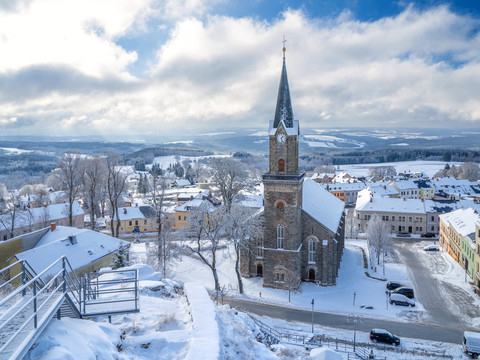 Schöneck_Blick vom Alten Söll Verschneite Aussicht auf eine Kirche mit spitzem Turm, umgeben von Winterlandschaft.