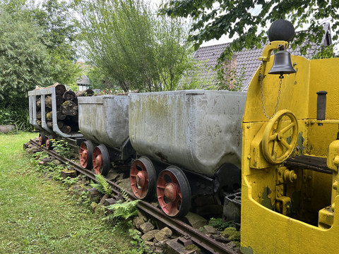 Bergisches Museum für Bergbau, Handwerk und Gewerbe Die alten Bergwerkswagen auf Schienen sind von Bäumen umgeben, eine gelbe Lok steht daneben.