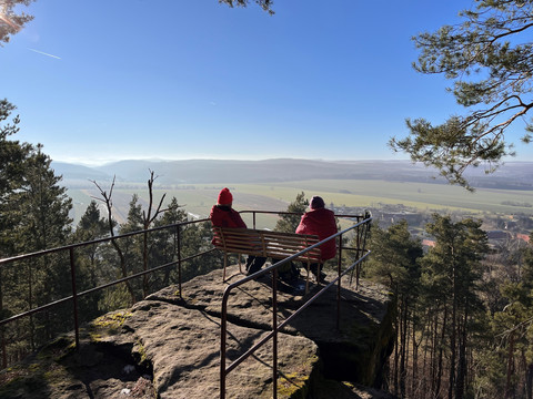 Zwei Personen in roten Jacken sitzen auf einer Bank auf einem Felsvorsprung, umgeben von Bäumen, mit Blick auf eine weite, sonnige Landschaft.Two people in red jackets are sitting on a bench on a rocky outcrop, surrounded by trees, with a view of a wide, sunny landscape.Dva lidé v červených bundách sedí na lavičce na skalnatém výběžku obklopeném stromy a mají výhled do širé, slunné krajiny.Dwie osoby w czerwonych kurtkach siedzą na ławce na skalistym występie, otoczonym drzewami, z widokiem na rozległy, słoneczny krajobraz.Twee mensen in rode jassen zitten op een bankje op een rotspunt, omringd door bomen, met uitzicht op een weids, zonnig landschap.Due persone in giacca rossa sono sedute su una panchina su uno sperone roccioso, circondato da alberi, con vista su un ampio paesaggio soleggiato.