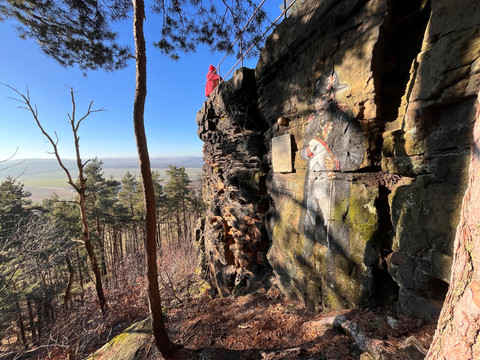 Person in rotem Mantel steht auf Felsvorsprung mit Blick auf bewaldete Landschaft; Felswand zeigt Napoleon-Graffiti und Gedenktafel.Person in red coat stands on rocky outcrop with view of wooded landscape; rock face shows Napoleon graffiti and memorial plaque.Osoba v červeném plášti stojí na skalním výběžku s výhledem na zalesněnou krajinu; na skalní stěně jsou vidět graffiti Napoleon a pamětní deska.Osoba w czerwonym płaszczu stoi na skale z widokiem na zalesiony krajobraz; na skale widać graffiti Napoleona i tablicę pamiątkową.Persoon in rode jas staat op rotspunt met uitzicht op bebost landschap; op de rots zijn graffiti van Napoleon en een gedenkplaat te zien.Persona in cappotto rosso in piedi su uno sperone roccioso con vista su un paesaggio boscoso; la parete rocciosa mostra graffiti su Napoleone e una targa commemorativa.