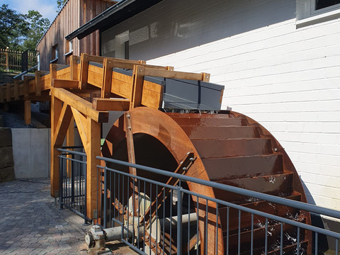 Dümmlinghauser Mühle Ein hölzernes Wasserrad dreht sich ruhig an einem modernen, weißen Gebäude unter klarem Himmel.