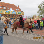 Aper Herbstmarkt „Mittelalterlicher Reiter in Kettenhaube auf einem Pferd beim Marktumzug, geführt durch historische Figuren, Publikum hinter nostalgischem Karussell ‚Böltes Pferde‑Karussell Anno 1904‘.“