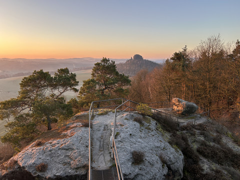 Kaiserkrone Steiniger Aussichtspunkt mit Geländer, umgeben von Bäumen, im Licht eines orangefarbenen Sonnenuntergangs, mit Hügeln im Hintergrund.