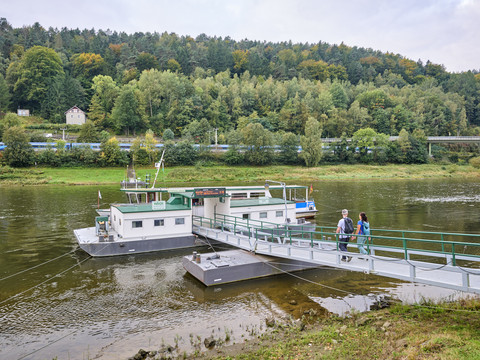 Ein Paar überquert eine grüne Brücke zu einer Fähre auf einem Fluss, umgeben von bewaldeten Hügeln und einem vorbeifahrenden Zug im Hintergrund.