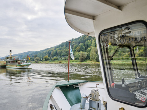 Fährüberfahrt Bad Schandau Blick von einem Boot auf einen Fluss mit einem Dampfer im Hintergrund, umgeben von bewaldeten Hügeln unter bewölktem Himmel.