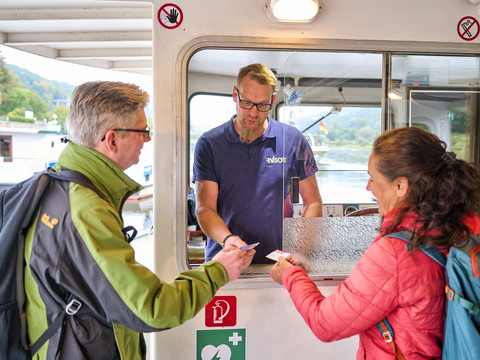 Ein Mann in grüner Jacke und eine Frau in roter Jacke kaufen Tickets bei einem Verkäufer in einem Bootsschalter; im Hintergrund ist Wasser zu sehen.