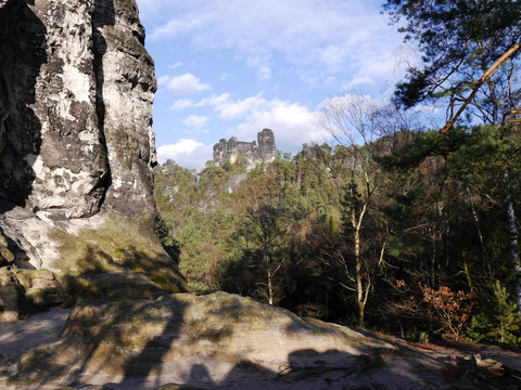 Felsformationen und bewaldete Hügel unter blauem Himmel mit Wolken; im Vordergrund ein großer, schattiger Felsen und Bäume.
