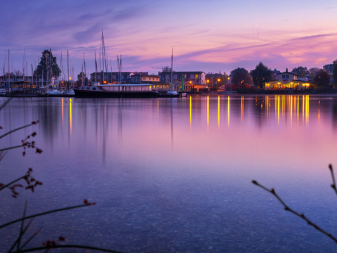 De roze en paars gekleurde lucht wordt weerspiegeld in het Cospudenmeer, een paar zeilboten liggen aangemeerd aan de pier en de sauna in het meer is helder verlicht, ontspanning, wellness, excursie, Leizpiger Neuseenland