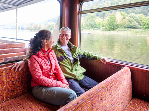 Ein Mann und eine Frau sitzen lächelnd auf einer Bootbank, er trägt eine grüne Jacke, sie eine rote, mit Blick auf einen Fluss und grüne Uferlandschaft.