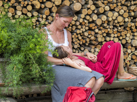 Eine Frau in einem weißen Top hält ein Kind in einem roten Kleid auf dem Schoß, vor einem Stapel Holzscheite, neben einem grünen Busch.A woman in a white top holds a child in a red dress on her lap, in front of a pile of logs, next to a green bush.Žena v bílém topu drží na klíně dítě v červených šatech před hromadou klád vedle zeleného keře.Kobieta w białej bluzce trzyma na kolanach dziecko w czerwonej sukience, przed stosem kłód, obok zielonego krzewu.Een vrouw in een wit topje houdt een kind in een rode jurk op haar schoot, voor een stapel boomstammen, naast een groene struik.Una donna con un top bianco tiene in grembo un bambino con un vestito rosso, davanti a una pila di tronchi, accanto a un cespuglio verde.