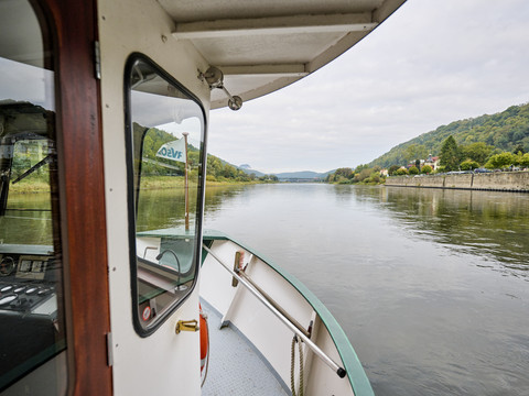 Blick von einer Fähre auf einen ruhigen Fluss, umgeben von bewaldeten Hügeln und einer Steinmauer am Ufer; das Wetter ist bewölkt.