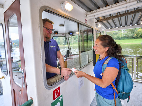 Eine Frau in blauem T-Shirt und Rucksack kauft ein Ticket bei einem Mann in einem Fährhäuschen. Beide lächeln, im Hintergrund ist Wasser zu sehen.