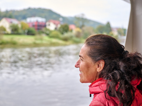 Fährüberfahrt Eine Frau in roter Jacke blickt lächelnd über einen Fluss; im Hintergrund sind unscharfe Häuser und grüne Hügel zu sehen.A woman in a red jacket looks out over a river with a smile; blurred houses and green hills can be seen in the background.Žena v červené bundě se s úsměvem dívá na řeku, v pozadí jsou vidět rozmazané domy a zelené kopce.Kobieta w czerwonej kurtce z uśmiechem spogląda na rzekę; w tle widać rozmyte domy i zielone wzgórza.Een vrouw in een rood jasje kijkt glimlachend uit over een rivier; op de achtergrond zijn vage huizen en groene heuvels te zien.Una donna con una giacca rossa si affaccia sorridente su un fiume; sullo sfondo si intravedono case sfocate e colline verdi.