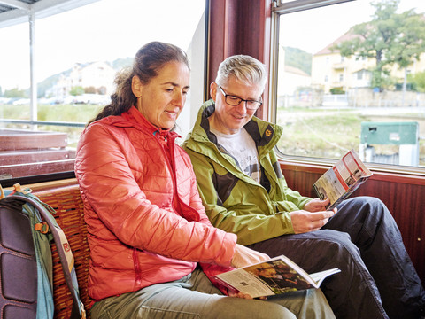 Ein Mann und eine Frau sitzen in einem Zugabteil, lesen Broschüren und tragen bunte Jacken; draußen ist eine ländliche Landschaft zu sehen.