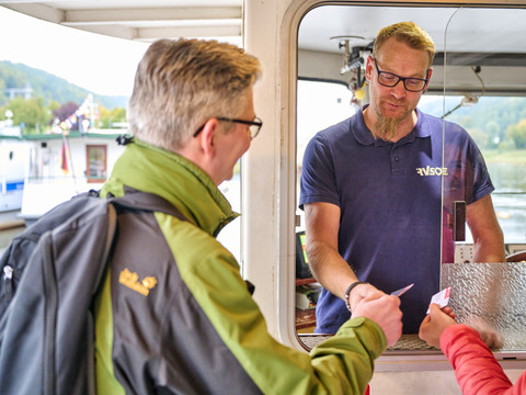 Im Gespräch mit dem Fährmann Ein Mann in grüner Jacke kauft ein Ticket bei einem Verkäufer in einem blauen Polohemd auf einem Boot. Im Hintergrund ist ein Fluss zu sehen.A man in a green jacket buys a ticket from a salesman in a blue polo shirt on a boat. A river can be seen in the background.Muž v zelené bundě si na lodi kupuje jízdenku od prodavače v modré polokošili. V pozadí je vidět řeka.Mężczyzna w zielonej kurtce kupuje bilet od sprzedawcy w niebieskiej koszulce polo na łodzi. W tle widać rzekę.Een man in een groene jas koopt een kaartje van een verkoper in een blauw poloshirt op een boot. Op de achtergrond is een rivier te zien.Un uomo con una giacca verde acquista un biglietto da un venditore con una polo blu su una barca. Sullo sfondo si vede un fiume.