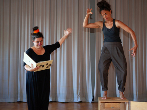 Two women in black clothing, one reading from a book, the other standing on a wooden box and gesticulating, in front of a gray curtain.