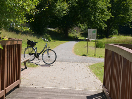 Symbolbild_Fahrrad_HD.JPG Geparktes Fahrrad an einer Holzbrücke. Im Grünen stehen Bäume und eine Infotafel.