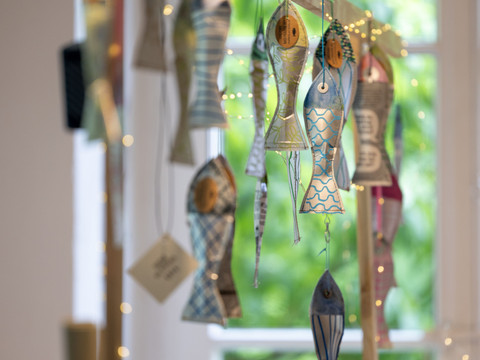 Papiermarkt 2024 Dekorative Papierfische und Lichterketten hängen an einem Holzgestell vor einem Fenster mit grünem Ausblick.