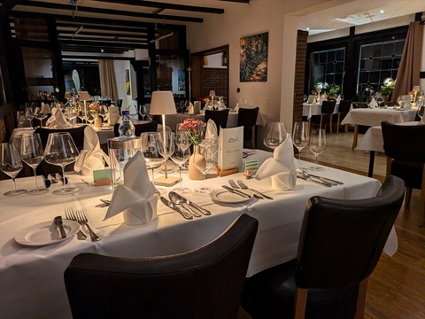 Eimermacher 01.jpg Festlich gedeckter Restaurant-Innenraum mit weiß gedeckten Tischen, eleganten Stühlen und gedämpfter Beleuchtung.