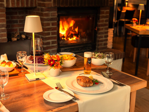 Eimermacher 03.jpg Gemütliches Restaurant mit gedecktem Tisch, leuchtendem Kamin im Hintergrund, Speisen und Getränken.