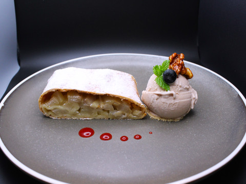 Eimermacher 09.JPG Ein Teller mit Apfelstrudel, einer Kugel Eis, einer Walnusshälfte und Beeren auf einem grauen Hintergrund.