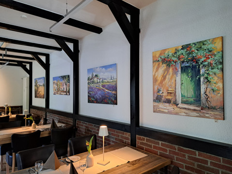 Eimermacher 16.jpg Gemütliches Restaurant mit sorgfältig gedeckten Tischen und Landschaftsgemälden an weißen Wänden.