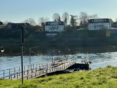 Ein Holzsteg führt über einen Fluss, im Hintergrund sind zwei mehrstöckige Gebäude und Bäume zu sehen, bei klarem Himmel und ruhiger Atmosphäre.