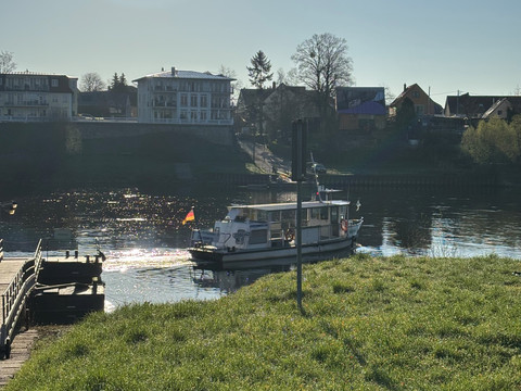 Fähre F10 Ein kleines Passagierschiff mit deutscher Flagge fährt auf einem Fluss, umgeben von grüner Wiese und Wohnhäusern im Hintergrund bei klarem Himmel.A small passenger ship flying the German flag sails on a river, surrounded by green meadows and houses in the background under a clear sky.Malá osobní loď plující pod německou vlajkou pluje po řece obklopené zelenou loukou a domy v pozadí pod jasnou oblohou.Mały statek pasażerski pod niemiecką banderą płynie po rzece, otoczony zieloną łąką i domami w tle pod czystym niebem.Een klein passagiersschip onder Duitse vlag vaart op een rivier, omringd door een groene weide en huizen op de achtergrond onder een heldere hemel.Una piccola nave passeggeri battente bandiera tedesca naviga su un fiume, circondata da un prato verde e da case sullo sfondo, sotto un cielo limpido.