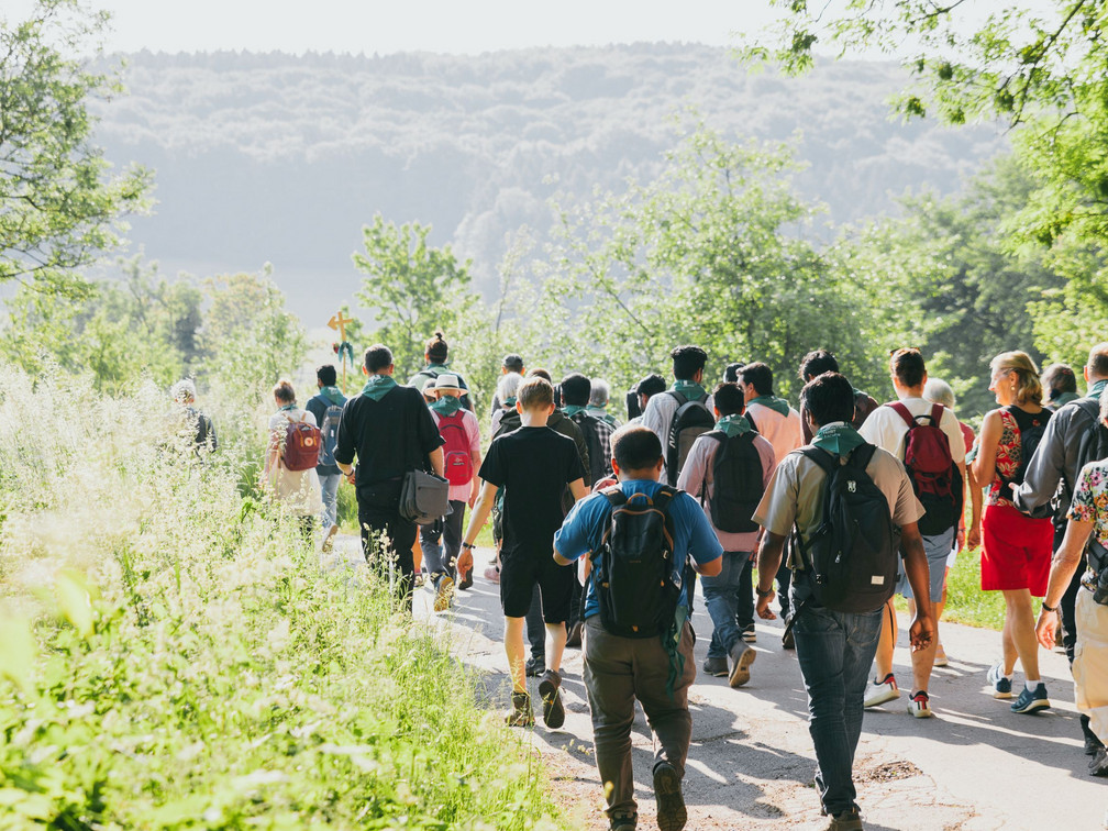 Pilgerwanderung (c) Domkapitel Aachen_Alexander Müller.jpg Eine Pilgergruppe wandert bei Sonnenschein auf einem grünen, von Bäumen gesäumten Weg. Menschen unterschiedlichen Alters und Geschlechts sind zu sehen. Sie tragen Rucksäcke und bequeme Kleidung, während sie sich auf ihrem spirituellen Pfad vorwärts bewegen.