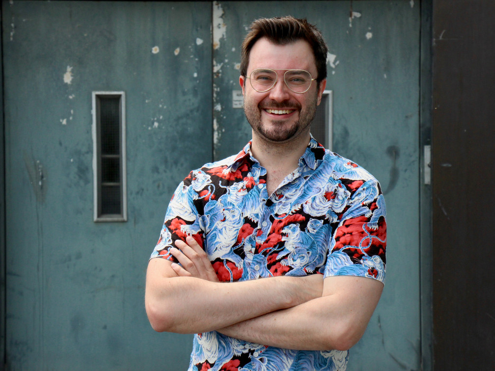 Klussmann.jpg Ein lächelnder Mann mit Brille und gemustertem Hemd vor einer blaugrauen industriellen Tür.