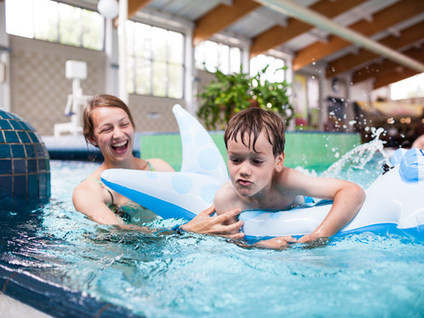 Erlebnisbad Frau und Kind spielen im Erlebnisbad auf einem blauen aufblasbaren Dino im Wasser.