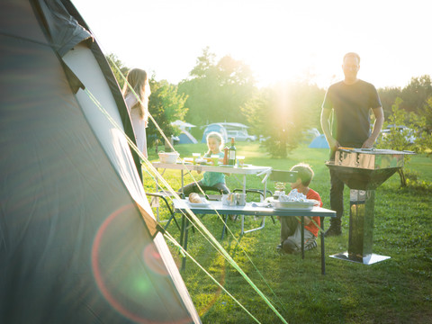Camping Familie beim Campen, genießt Mahlzeit im Freien, Sonnenuntergang im Hintergrund, Zelt rechts.