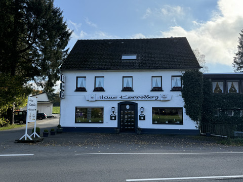 Haus Koppelberg Außenansicht Gasthaus mit weißen Wänden, dunklem Dach und grüner Rankenverzierung, daneben eine Straße.