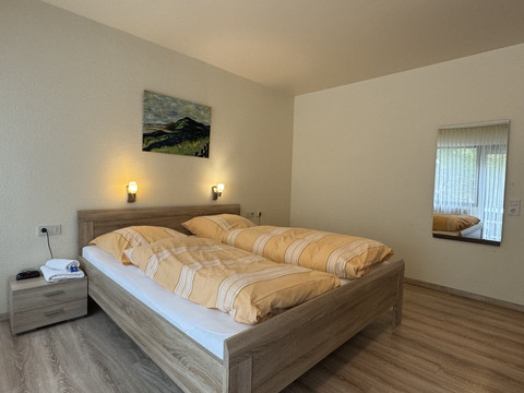 Innenansicht Doppelzimmer Haus Koppelberg Helles Schlafzimmer mit Doppelbett in Holzoptik, gelber Bettwäsche, Wandbild und Fensterspiegel.