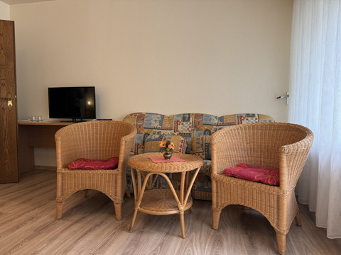 Innenansicht Doppelzimmer Haus Koppelberg Gemütliche Sitzecke mit zwei Rattansesseln und Tisch, dahinter ein buntes Sofa und ein TV im Hintergrund.