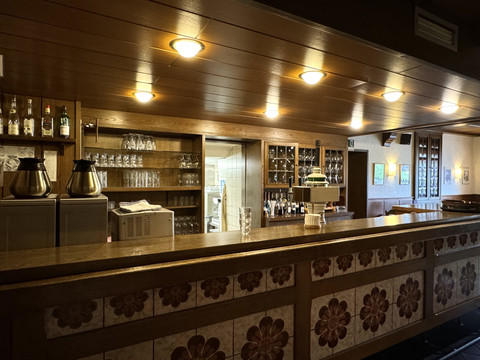 Ansicht Bar - Haus Koppelberg Holzvertäfelte Bar mit gemusterten Fliesen, Gläsern im Regal, gedämpfter Beleuchtung und Thekeneinrichtung.