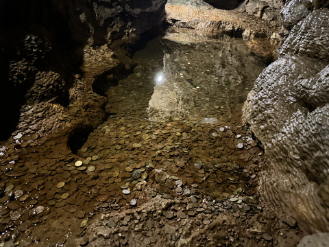 Ansicht Tropfsteinhöhle Ein unterirdischer Höhlensee mit stiller Wasseroberfläche, von Münzen bedeckt, umgeben von Felsen.