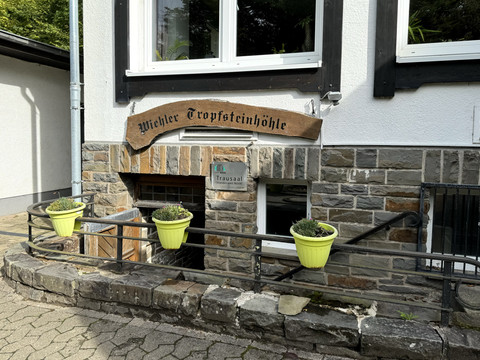 Ansicht Eingang zur Tropfsteinhöhle Schild "Wiehler Tropfsteinhöhle" an steinverkleideter Hauswand, flankiert von drei Blumenkübeln.